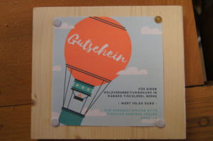 mit Heftzwecken auf einem Brett befestigtes Blatt zeigt einen Heißluftballon. Text: Gutschein über einen Kurs in Mannes Tischlerei.